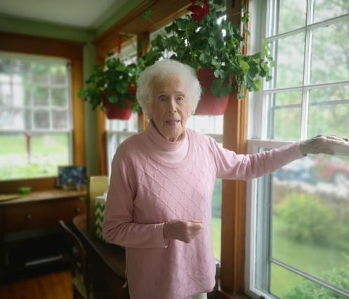 An image of 101 year old Joy Saunders standing in her home by her window, she is wearing a pink sweater. 