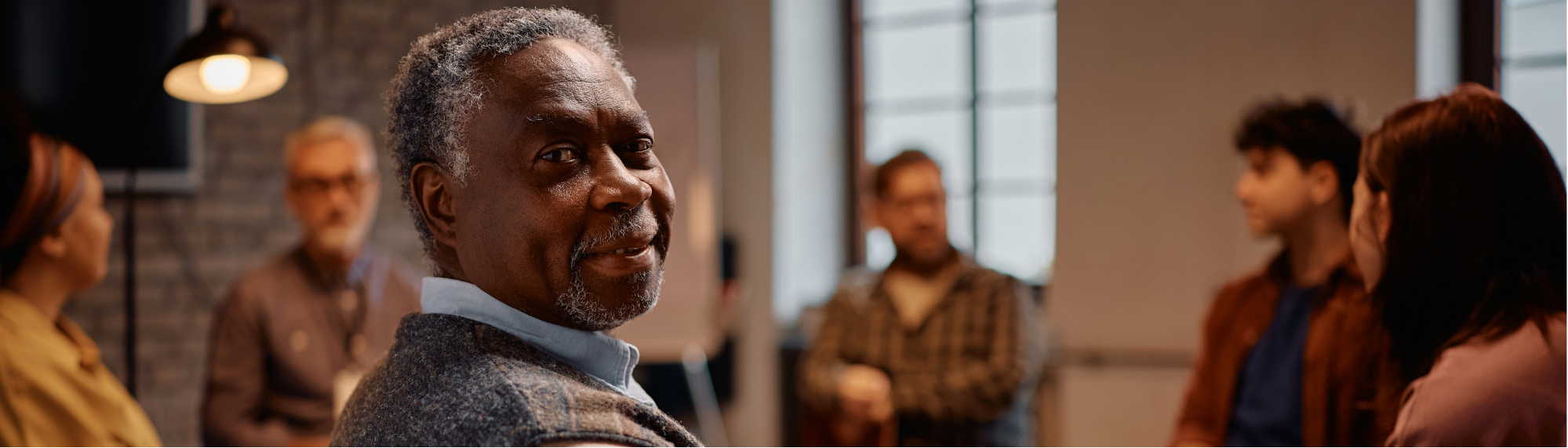Group of adults sitting in a dimly lit room in a circle. One older adult man is in focus looking at the camera. 
