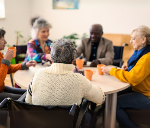 Grouds of 5 elder people around a circle table, having a conversation, orange mugs infront of them