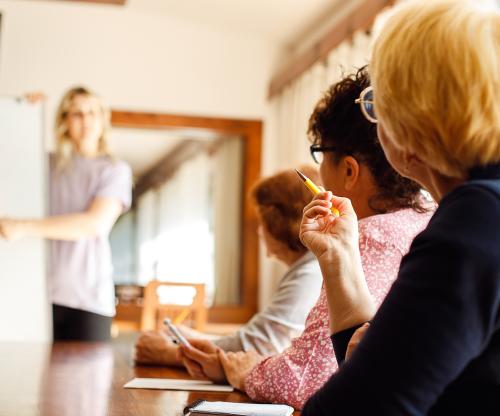 Three women seated at a boardroom table looking toward a blonde haired woman standing at the head of the table and pointing to something on a paper presentation,. The women standing and presentation is out of focus.